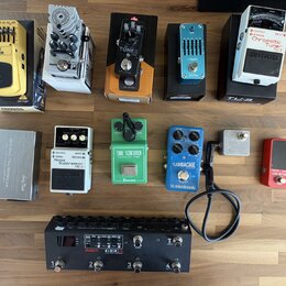 Sestava efektů pro kytaru rozložená na dřevěném povrchu. V horní řadě jsou pedály Behringer GRAPHIC EQUALIZER EQ700, AMPLITUBE, TONEX, S-BAND a BOSS…