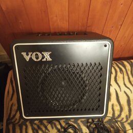 Černý kytarový kombo zesilovač s bílým rámečkem a mřížkou z kovového pletiva. Na přední straně je umístěno stříbrné logo VOX. Před přístrojem leží če…