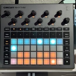 Šedý sampler Novation Circuit Rhythm s mřížkou podsvícených padů v modrých a oranžových barvách. V horní části jsou umístěny ovládací knoflíky s popi…