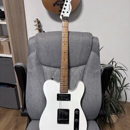 Bílá elektrická kytara s hnědým hmatníkem, opřená o šedé kancelářské křeslo. Na hlavě hmatníku je černý nápis Squier Telecaster. Kytara má jeden sním…