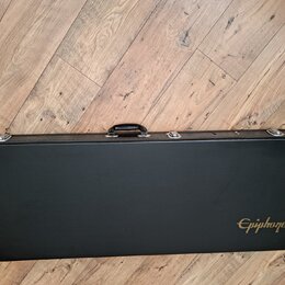 Černý tvrdý kufýr na hudební nástroj s rukojetí a kovovými zapínacími prvky. V pravém dolním rohu je zlatý nápis Epiphone. Povrch kufýru vykazuje dro…