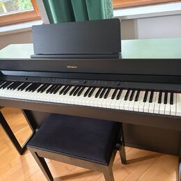 Černý digitální piano v podobě konzole s přiloženým polstrovaným černým stolečkem. Na přední horní hraně je umístěn nápis Roland. Nástroj má standard…