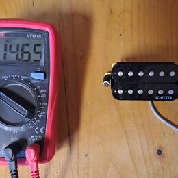 Černý humbucker s dvěma řadami bílých pólů a nápisem SCHECTER na spodní části. Snímač je připojen šedým kabelem a v pozadí je červený digitální multi…
