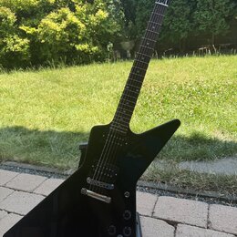 Černá elektrická kytara s ostře hranatým tělem typu Explorer umístěná na černém stojanu. Na hlavě hmatníku je bílý nápis Gibson. Kytara má dva humbuc…
