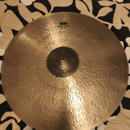 Zlatavý kovový činel s viditelným kovánívým povrchem a klepaným středem. V horní části je černý nápis HH, pod ním text Rare Bell Dry Ride a dále 21"/…
