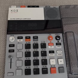 Šedý sampler s černým displejem a ovládacími prvky. Na horní bílé části je nápis K.O. II, pod ním japonské znaky サンプラー a text 64 MB SAMPLER COMPOSER.…