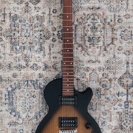 Elektrická kytara s černým korpusem v přechodovém odstínu do hnědého (sunburst) a tmavým hnedým hmatníkem. Na hlavě je bílý nápis Epiphone a vpravo p…