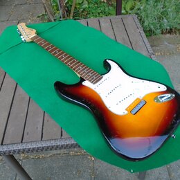 Elektrická kytara s trupem v barevném přechodu od červené přes oranžovou k žluté a bílým pickguardem. Na hlavě hmatníku je nápis Fender Stratocaster.…