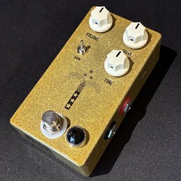 Zlatý efekt pedál s kovovým povrchem a třemi bílými oválnými knoflíky. Na horní straně jsou nápisy VOLUME, DRIVE a TONE, přepínač s popiskem GAIN a g…