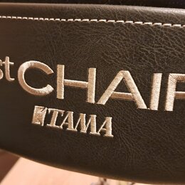 Detail černé polstrované sedací plochy z materiálu imitující kůži s bílým prošíváním. Na povrchu je vyšívka v bílé barvě s nápisy 1st CHAIR a TAMA. P…