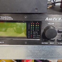 Černý rackový procesor s nápisy FRACTAL AUDIO SYSTEMS a Axe-Fx II PREAMP/ FX PROCESSOR. Zařízení má zelený displej, ovládací knoflík VALUE a řadu tla…