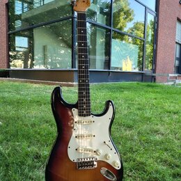 Elektrická kytara s tmelem v barvě sunburst, bílým pickguardem a hnedým hmatníkem. Na hlavě je viditelné logo Fender. Nástroj má tři snímače, pětipos…