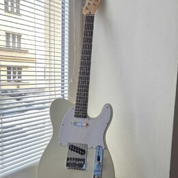 Světle šedá elektrická kytara s bílým pickguardem a hnedým hmatníkem. Na hlavě je nápis Squier by Fender TELECASTER. Nástroj vykazuje známky běžného…