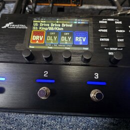 Černý kytarový procesor s nápisy FRACTAL AUDIO SYSTEMS a VP4 VIRTUAL PEDALBOARD. Zařízení má digitální displej s texty UI: Drive Drive Drive!, S1: Ki…