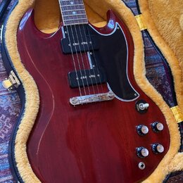 Elektrokytara s tmavě červeným lakovaným povrchem a černým pickguardem v hnědém plišovém kufru. Nástroj má dva humbuckery, stříbrný most a čtyři ovlá…
