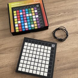 Na fotografii je černý MIDI kontrolér Novation Launchpad X s mřížkou bílých podsvícených polí a ovládacími tlačítky po stranách. Vedle zařízení leží…