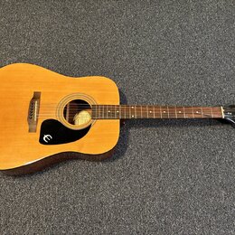Akustická kytara s přírodním dřevem, tmavě hnědými boky a černým plechtřem. Na hlavě hmatníku je bílý nápis Epiphone. Uvnitř ozvučovacího otvoru je v…