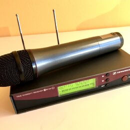 Bezdrátový mikrofon s černou mřížkou a stříbrno-šedým tělem leží na černém přijímači. Na přijímači je nápis TRUE DIVERSITY RECEIVER ew 100 G2, logo a…