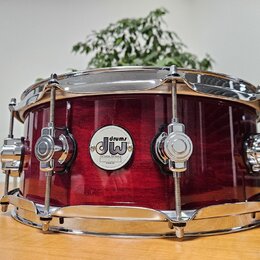 Červený snare drum s kovovým obsazem a lesklým povrchem. Uprostřed korpusu je bílý oválný štítek s nápisy drums, dw a DESIGN SERIES. Nástroj vykazuje…
