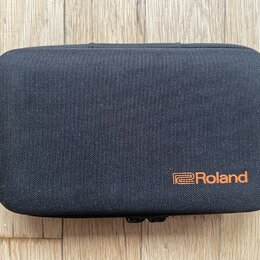 Černý obdélníkový pouzdro z textilie s texturovaným povrchem. V pravém dolním rohu je oranžové logo a nápis Roland. Pouzdro leží na dřevěném podkladu.