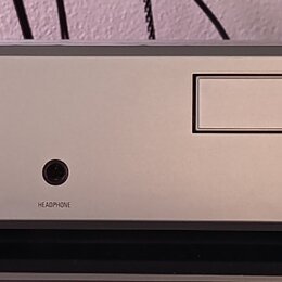 Stříbrný audio komponent s digitálním displejem, na kterém svítí zelený nápis AM 7.28. Na pravé straně je logo v kruhu a nápis LINN, v dolní části je…