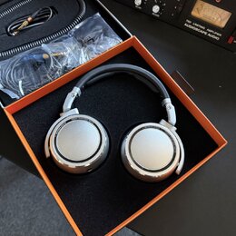 Stříbrná studiová sluchátka s černým náušníkem v oranžovo-černé krabici. Na obou mušlích je nápis STUDIO HEADPHONE 600 a BEHMANN BERLIN. Vedle sluchá…