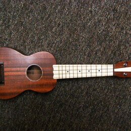Hnědý dřevěný ukulele s hnedým korpusem a světlým hmatníkem. Uprostřed ozvučovacího otvoru je vidět štítek s nápisem "Kala". Nástroj vykazuje známky…