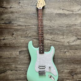 Světle zelená elektrická kytara typu Stratocaster s bílým perleťovým pickguardem a hnedým hmatníkem. Na hlavě je černý nápis Fender Stratocaster. Nás…