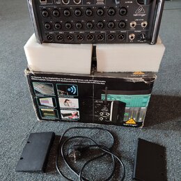 Digitální mixážní pult Behringer X AIR XR18 v černé barvě, umístěný na bílém polystyrenovém podkladu. Vedle přístroje je originální kartonová krabice…