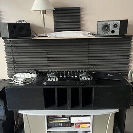 Na fotografii je set DJ vybavení umístěné na černých akustických panelech a bílém stolku. V horní části jsou dva černé studiové monitory, vprostřed č…