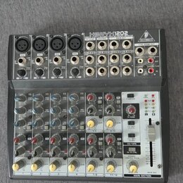 Šedý mixovací pult s nápisem XENYX 1202 a logem BEHRINGER. Zařízení obsahuje čtyři mikrofonní vstupy XLR, několik lineárních vstupů RCA a jack, poten…