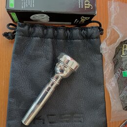 Stříbrný moutur z kovu položený na černém koženém vak s vyraženým nápisem KOSS. Nad nimi je černá krabička s bílými nápisy Bach Genuine Mouthpiece a…