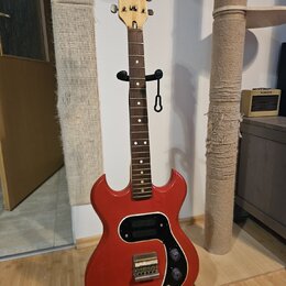 Červená elektrická kytara s dřevěným hmatníkem a světle hnědým hlavním hmatníkem. Na hlavě je nápis "disco plena". Tělo má černý pickguard, dva ováln…