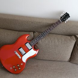 Elektrická kytara s červeným lakovaným korpusem a hnědým hmatníkem. Nástroj má dva humbuckery, bílý plech sylovací kryt a čtyři zlaté ovládací knoflí…