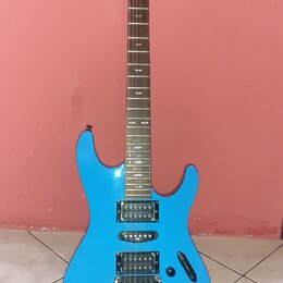 Modrá elektrická kytara s hrazdicem a dvěma humbuckery v pozici kobylníku a krku. Na hlavě hmatníku je bílý nápis Ibanez. Nástroj stojí na černém sto…