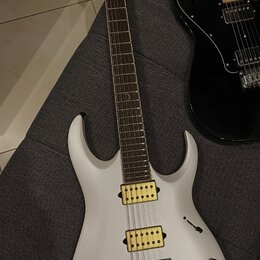Bílá elektrická kytara s matným povrchem a hnědým hmatníkem. Na hlavě je nápis Ibanez, na těle jsou dva žluté humbuckery a černý kobylník. Vedle ní l…