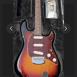Elektrická kytara s trupem v barevnosti sunburst a tmavě červeným pickguardem s bílými vlożkami. Nástroj leží v černém polstvu, vedle něj jsou přilož…