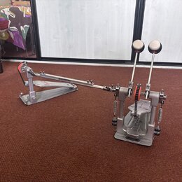 Dvojpedál na bicí v šedé barvě s kovovým povrchem. Na základní desce pedálu je viditelné červené logo. Mechanismus obsahuje řetězy, pružiny a dvě šla…