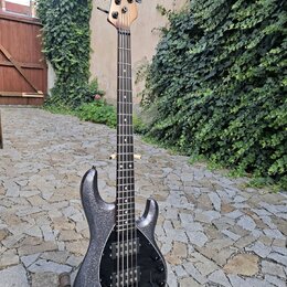 Elektrická basová kytara s tmavě šedým trpytivým povrchem těla a hnedým dřevěným hmatníkem a hlavou. Na hlavě je svisle napsaný nápis Ibanez. Tělo má…
