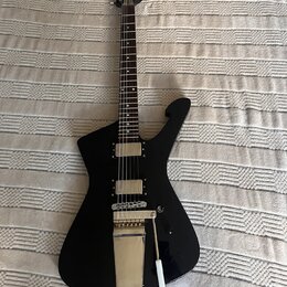 Černá elektrická kytara s atypickým, ostře hranatým tvarem těla a tmavým hmatníkem. Nástroj má dva humbuckery s kovovými kryty a stříbrný tremolo sys…