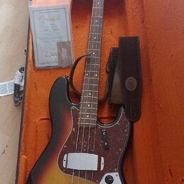 Elektrická basová kytara s trupem v barevnosti sunburst a hnědým hmatníkem, umístěná v otevřeném kufru s oranžovou výstuhou. Na hlavě je nápis Fender…