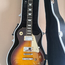 Elektrická kytara typu Les Paul s barevným přechodem od žluté k tmavě hnědé v hardcase s černým vnitřkem. Na hlavě je nápis "Vantage". Kytara má dva…