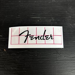 Bílý obdélníkový štítek s černým nápisem Fender v kurzívě. Povrch je proříznut červenou mřížkou s tenkými čarami. Štítek je umístěn na tmavém, textur…