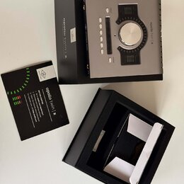 Stříbrné audio rozhraní s černým ovládacím panelem a velkým centrálním knoflíkem, umístěné v černé krabici. Na zařízení a krabici je uveden nápis apo…