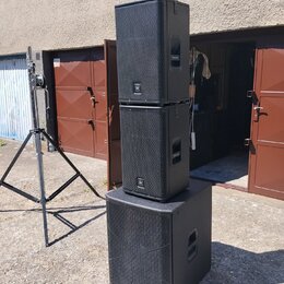 Tři černé reproduktorové bedny v kostce s kovovou mřížkou, naskládané na sebe. Na každém reproduktoru je v dolní části viditelné bílé logo znázornují…