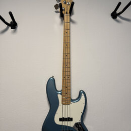 Modrá elektrická basová kytara s bílým pickguardem a světle hnědým hmatníkem. Na hlavě je vidět logo "Fender" a nápis "Made in Japan". Nástroj vykazu…