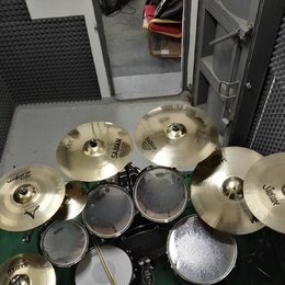 Sada cymbálů a bubnů v zeleném studiu. Na cymbálech jsou viditelné nápisy SABIAN, Zildjian a Soultone. Bubny mají bílé koženky s logem Evans. Povrchy…