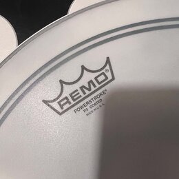 Detailní záběr na bílou, potaženou bicí koženu. Na povrchu je černý nápis REMO s logem koruny a text POWERSTROKE® P3 COATED MADE IN U.S.A. Kožena je…