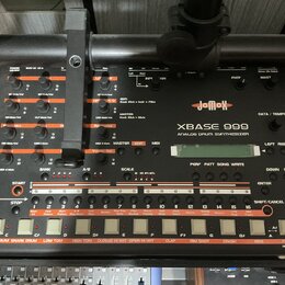 Šedý analogový drum synthesizer značky jomox, model XBASE 999. Zařízení má mnoho ovládacích knoflíků, posuvníků a bílá tlačítka v dolní části s popis…