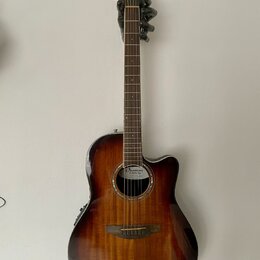 Akustická kytara s cutawayem v tmavě hnědém sunburst provedení. Na hmatníku a hlavě není vidět žádný nápis, ale na ozvukové desce je v rámci rozetové…